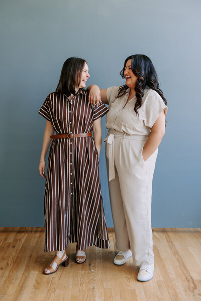 Bridget and Vanessa smile at each other in front of gray-blue wall while Vanessa has one hand in her pocket and the other resting on Bridget's shoulder.