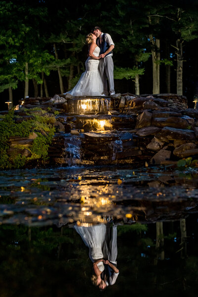 night portraits of a wedding couple at a wedding venue, captured by their Austin wedding photographer. Their reflection is in the lake water while they bow their heads together.