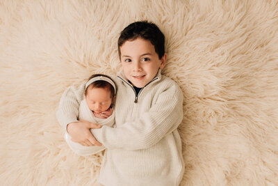 A boy smiling at the camera, holding his newborn baby sister at her newborn session