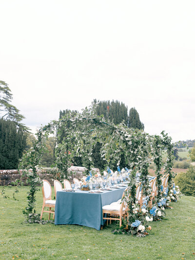Outdoor reception table with blue tablecloths, floral centerpieces, greenery arches, and floral arrangements on the ground