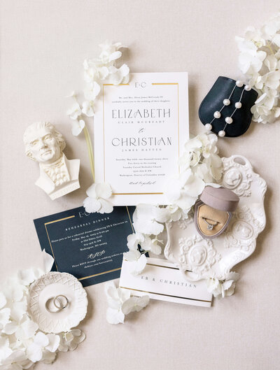 A flat lay of elegant white and gold wedding stationery featuring invitations, accompanied by white flowers, a small bust statue of a president, jewelry, rings, and an RSVP card on a light background.