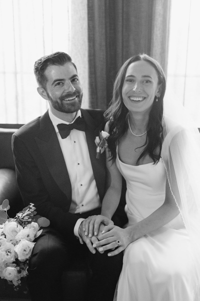 Black and white image of newly wed couple sat, holding hands and smiling at the camera