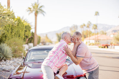 Engaged couple sharing a kiss on the hood of a red vintage car in Palm Springs, framed by desert plants, palm trees, and mountain views.