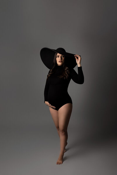 Expectant London, Ontario Mom at a maternity photoshoot in top photography studio. Mom is earing a black collared bodysuit, bare legs, and a wide brimmed felt hat. One had is under her bump, the other hand is gently touching her hat. She is looking at the camera with a half smile.