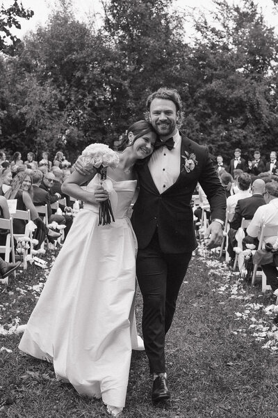 The bride playfully runs away as the groom chases her. She is dressed in a mid length satin white dress and has a short veil flowing behind her.
