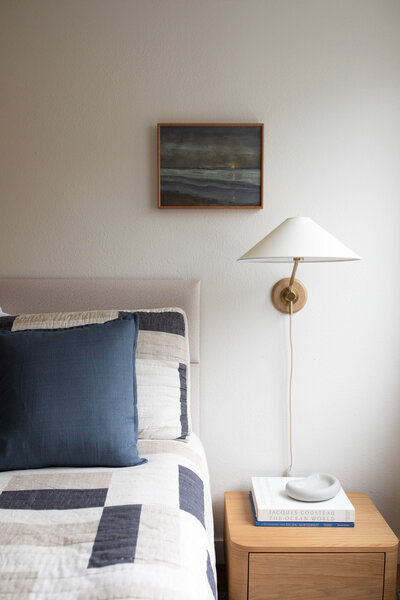 A bed with a nightstand to the right and a lamp and painting above it. Representing a beach house Interior Design by Winsome Home Interiors.