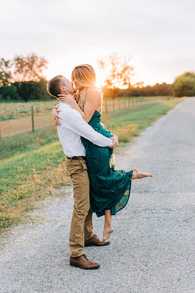 Man in white shirt and khaki pants picking up woman in green maxi dress with sun setting behind them