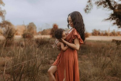 A woman in an orange dress smiles at a young child in a grassy outdoor field with trees, showcasing a touching moment by a Macon motherhood photographer.