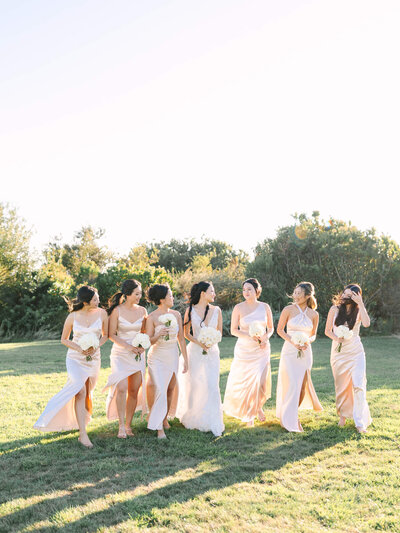 Seven women in pastel dresses walk on grass, smiling and holding bouquets. The sky is clear, and greenery surrounds them.