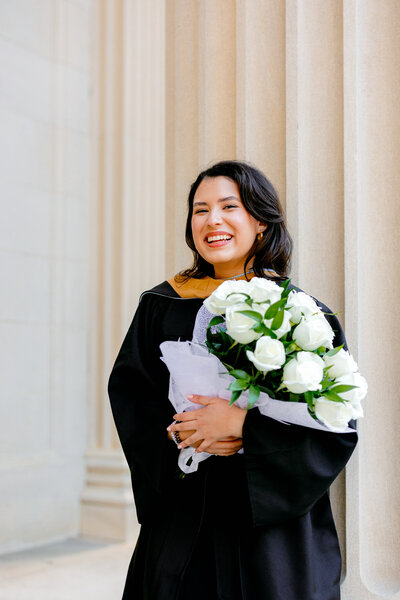 UNC graduate in black gown and hood holding flowers while leaning against the columns of Wilson Library.