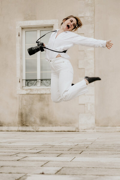 Laura, photographe sur un mariage en Vendée, tenue blanche, saute de joie, appareil photo en main, devant un bâtiment en arrière-plan.