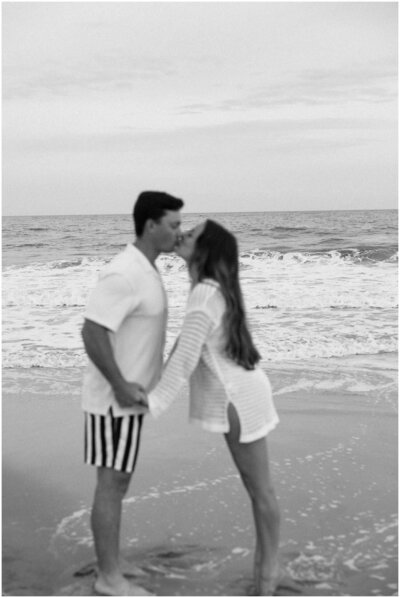 A happy couple in a black and white image kissing and holding hands on a beach in white shirts during their engagement session