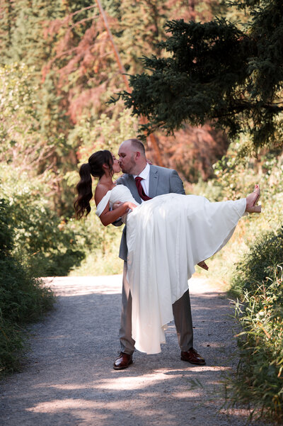 groom holding bride in his arms kisses her on a pathway in the forest