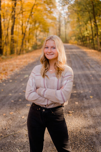 Teenage girl poses for the camera outside standing in the middle of a backroad with fall leaves.
