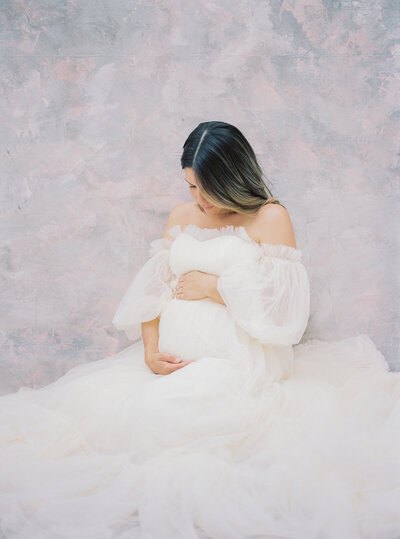 A pregnant mother cradles her belly while wearing a long white gown during her Maryland maternity session in studio