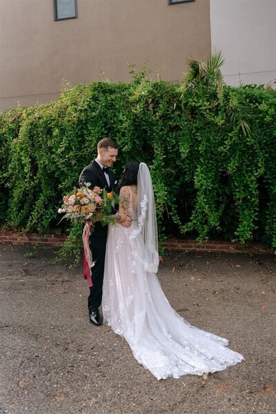 A newlywed couple shares a tender moment outdoors at dusk, with the groom dressed in a gray vest and white shirt, embracing the bride who is radiant in an off-the-shoulder lace wedding gown. They are smiling joyfully, with the groom leaning in for a kiss on the bride's forehead. The backdrop features a dramatic sunset sky in warm hues, with silhouettes of mountains and trees, adding to the romantic ambiance of the scene.
