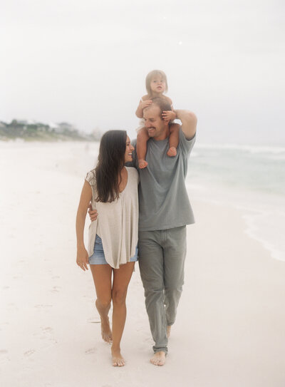 A mother and father walking on the beach in destin, florida with their little girl with blonde hair riding on the dad's shoulders.