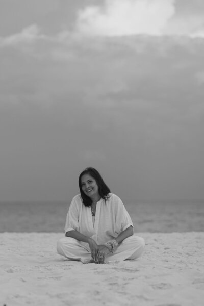 Black and white image of woman in white pants and white shirt with dark brown shoulder length hair sitting on the beach with the ocean behind her she is looking directly at the camera smiling