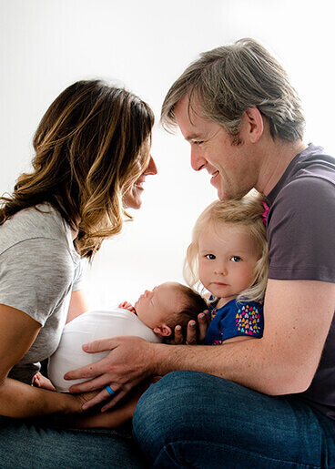 A family of four with a toddler girl and a newborn girl. Mom and dad are smiling at each other and the toddler is looking at the camera. They are all holding the newborn.