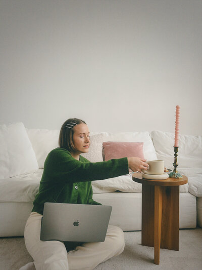 Margarita sits on a whit carpeted floor, reaching for her cream-coloured teacup, her laptop in her lap.