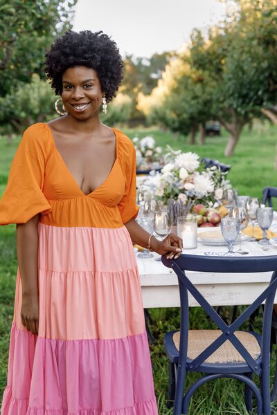 Fallon, smiling in an orange and pink dress, in front of an outdoor table setup at Breeze Hill Farm