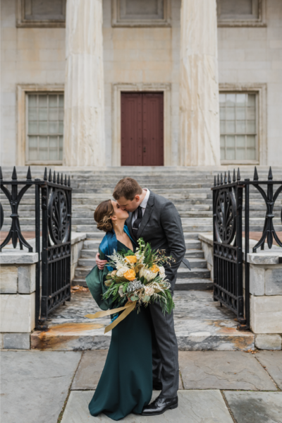 Couple in a green gown and gray suit kissing outdoors in front of a building.