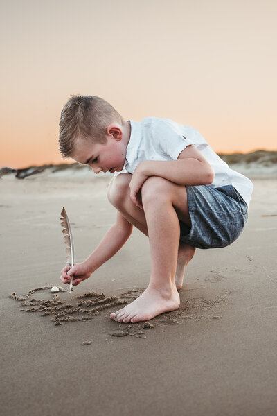YOUNG BOY KNEELING ON THE BEACH WRITING IN THE SAND WITH A BIRD FEATHER IN PORT ARANSAS TEXAS AT SUNSET