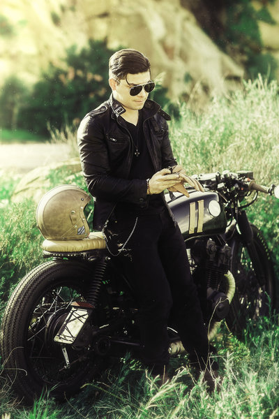 Man in all black with leather jacket and sunglasses leaning against motorcycle in the hills of Los Angeles