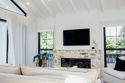 Bright living room with a stone fireplace, wall-mounted TV, and large windows. Neutral-toned furniture and plants showcase bespoke interior design, epitomizing luxury home design in Newcastle NSW.