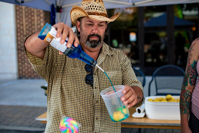 A man wearing a straw cowboy hat and checkered shirt pouring liquid from a bottle into a clear container at what looks like an outdoor event or market. He has a beard and is focused on the pouring action.