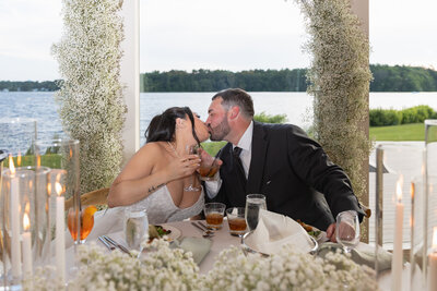 The Lakehouse Inn MA  | Kelsey Sheehan Photography Timeless Rhode Island Weddings | A bride and groom in formal attire share a kiss at a table overlooking a lake. They each hold a drink, surrounded by flowers, creating a joyful atmosphere.