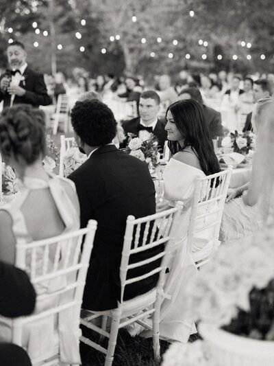 Black and white photo of wedding guests seated outdoors under string lights, listening to speeches during the reception.