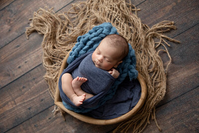 Newborn baby swaddled in a soft blue wrap, resting in a wooden bowl with a chunky knit blue blanket and textured burlap backdrop. Cozy and professional newborn photography setup.