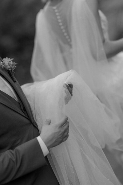 Black and white close-up of a groom gently holding the bride’s delicate tulle gown during their wedding at The Garden Villa in Winter Park, Florida — a timeless moment of love and support.