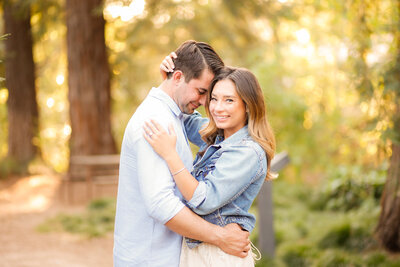 Engaged girl looks at camera as her fiancé snuggles his head into the side of her forehead and holds her lovingly with greenery in the background. Photo taken at UC Davis arboretum by wedding photographer Philippe Studio pro near sacramento.