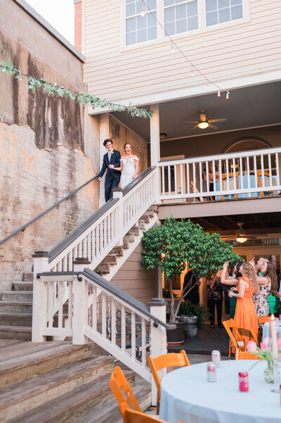 A bride and groom walk down a wooden staircase at an outdoor wedding venue, with guests seated and taking photos below. The space is decorated with greenery and warm string lights.