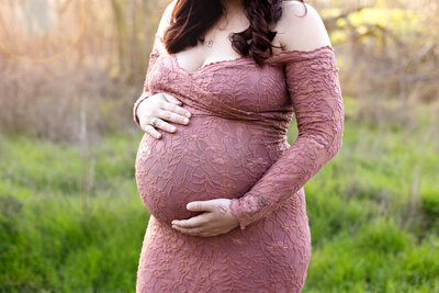 A pregnant woman cradles her belly and is wearing a pink lacy dress