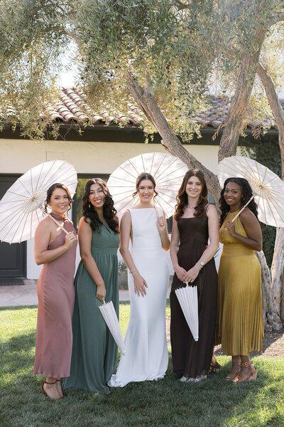 Bride and bridesmaids holding paper parasols, dressed in earthy-toned gowns at an outdoor wedding. Styled by a Central Coast wedding planner for a timeless celebration.