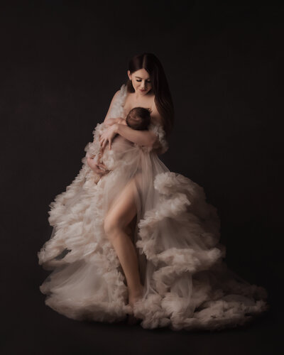 holding baby boy posed in studio setting wearing a fluffy couture gown on black near edgewood