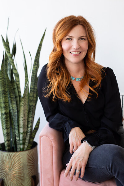 woman sitting on the floor, smiling and touching her head, looking at the camera. Photo taken in a studio where there are pampas grass hanging from the ceiling