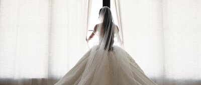 Details of the dress of a bride looking out a window at her wedding with a veil on.