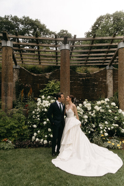 Bride and groom by white hydrangeas under the pergola at The Crane Estate in Ipswich, Massachusetts.