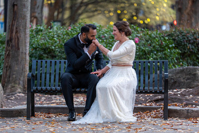 A newlywed couple sitting on a bench while one kisses the other's hand 