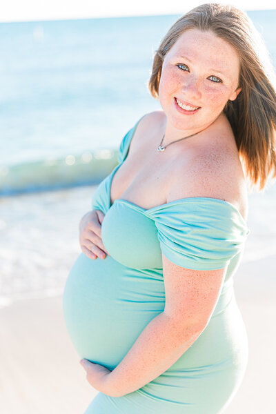 mother to be in a long teal dress on the beach cradling her baby bump