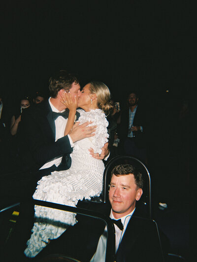 Flash film photo of bride and groom kissing while sitting on the back of their getaway car