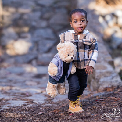 "A young boy smiles while holding a teddy bear during an outdoor portrait session by a Simpsonville photographer."