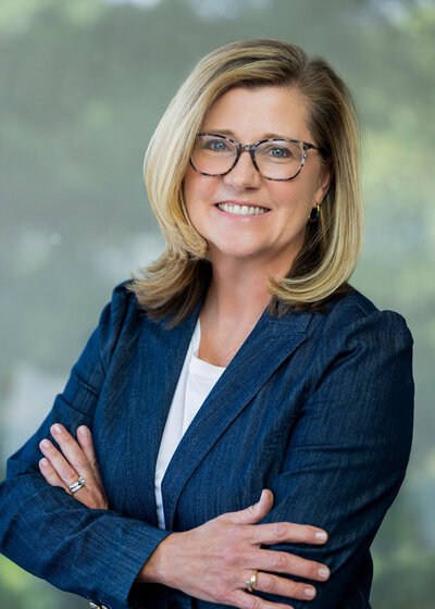Woman in a dark blue blazer with blonde hair and glasses smiling against a green background