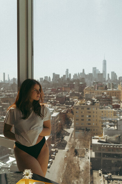 Woman looks out window and lifts shirt, directed by NYC boudoir photographer Elsie Goodman.