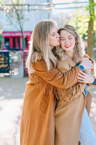 Lesbian couple embracing on rue de l’Abreuvoir in Montmartre, with one woman kissing the other’s cheek during a romantic Paris couples photoshoot.