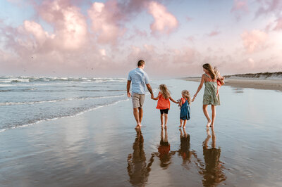 A mother and father walk along the beach in Corpus Christi, Texas, with their three young daughters while holding hands and looking at each other.